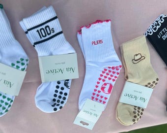 May include: Five pairs of athletic socks with various designs and text. The socks are white, tan, and black, with details like hearts, a cowboy hat, and the words "100s", "Pilates", and "Little But". Each pair has a tag that says "Aia Active".