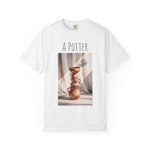 Puede incluir: Camiseta blanca con el texto "A POTTER" sobre una ilustración en acuarela. La ilustración representa una pila de cuencos de cerámica marrones y una ramita de lavanda. La camiseta tiene cuello redondo y mangas cortas.