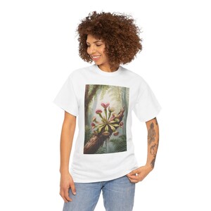 Puede incluir: Camiseta blanca con una ilustración detallada de una planta Venus atrapamoscas en una rama de árbol. La obra de arte muestra las características distintivas de la planta en rojo y verde, sobre un fondo de bosque. La camiseta la lleva una persona.