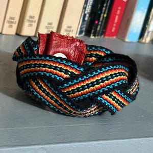 Peut inclure: Nœud décoratif tissé avec des fils noirs, bleus, oranges et jaunes. Un petit objet rouge est placé à l'intérieur du nœud. Le nœud repose sur une surface grise, avec un arrière-plan flou de livres.