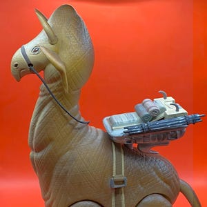 Op de afbeelding: Een bruin Bantha-speelgoed met een zadel en een gemonteerd wapen. Het speelgoed heeft een gedetailleerd ontwerp, inclusief een textuurhuid en een harnas. Het wapen is grijs en bruin, en het speelgoed staat tegen een rode achtergrond.