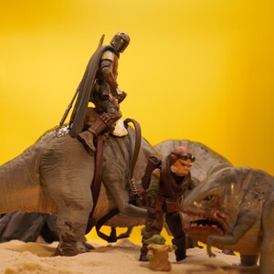Puede incluir: Un juguete de dinosaurio marr&oacute;n y gris con una figura de un Mandaloriano montando en su espalda. Un segundo juguete de dinosaurio m&aacute;s peque&ntilde;o con una figura de un personaje de Star Wars est&aacute; en primer plano.