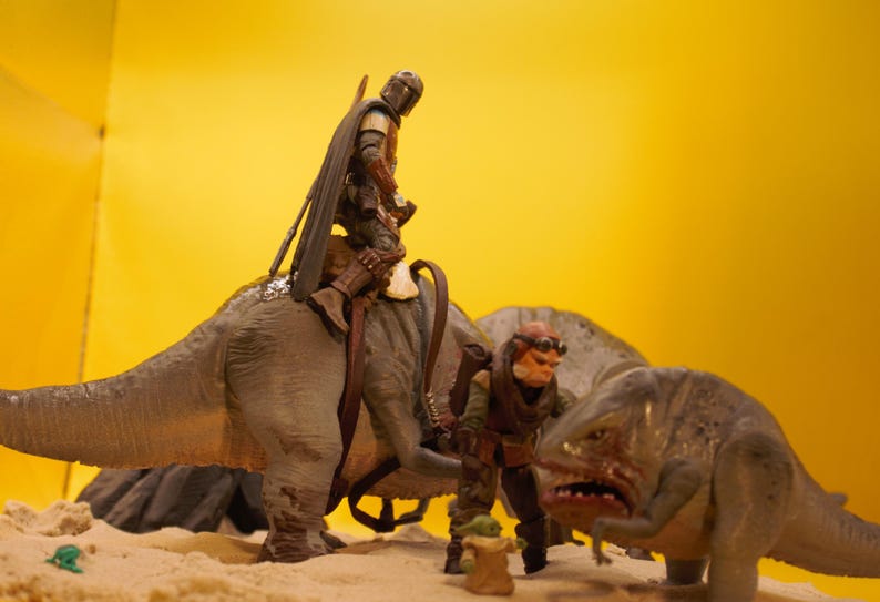 Puede incluir: Un juguete de dinosaurio marr&oacute;n y gris con una figura de un Mandaloriano montando en su espalda. Un segundo juguete de dinosaurio m&aacute;s peque&ntilde;o con una figura de un personaje de Star Wars est&aacute; en primer plano.