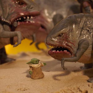 Puede incluir: Una peque&ntilde;a figura verde y marr&oacute;n se encuentra entre dos grandes criaturas reptilianas grises. Las criaturas tienen dientes afilados y est&aacute;n en una pose amenazante.