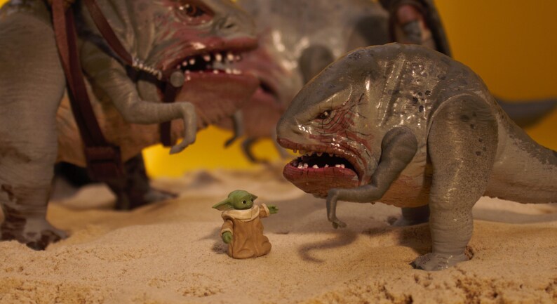 Puede incluir: Una peque&ntilde;a figura verde y marr&oacute;n se encuentra entre dos grandes criaturas reptilianas grises. Las criaturas tienen dientes afilados y est&aacute;n en una pose amenazante.
