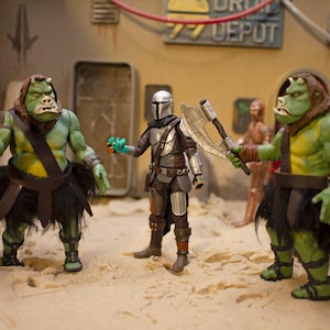 K&ouml;nnte beinhalten: Drei Actionfiguren, zwei gr&uuml;ne und eine silberne, stehen auf einer sandigen Oberfl&auml;che. Die gr&uuml;nen Figuren sind humanoid mit gro&szlig;en K&ouml;pfen und tragen schwarze R&ouml;cke. Die silberne Figur ist ein Mandalorianer mit einem Helm und einer silbernen Axt. Der Hintergrund ist ein W&uuml;sten-Set mit einem Schild, auf dem "DRIV DEPOT" steht.