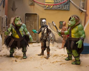 Könnte beinhalten: Drei Actionfiguren, zwei grüne und eine silberne, stehen auf einer sandigen Oberfläche. Die grünen Figuren sind humanoid mit großen Köpfen und tragen schwarze Röcke. Die silberne Figur ist ein Mandalorianer mit einem Helm und einer silbernen Axt. Der Hintergrund ist ein Wüsten-Set mit einem Schild, auf dem "DRIV DEPOT" steht.