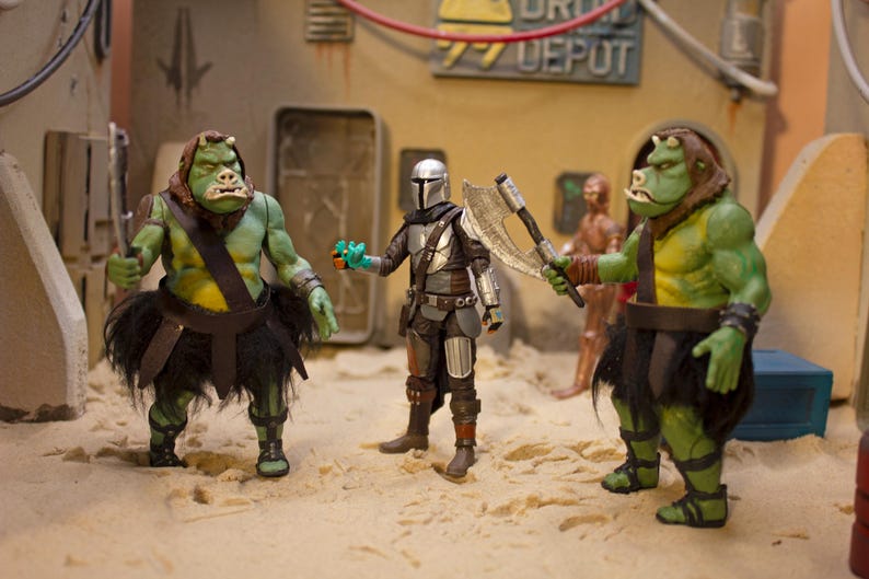 K&ouml;nnte beinhalten: Drei Actionfiguren, zwei gr&uuml;ne und eine silberne, stehen auf einer sandigen Oberfl&auml;che. Die gr&uuml;nen Figuren sind humanoid mit gro&szlig;en K&ouml;pfen und tragen schwarze R&ouml;cke. Die silberne Figur ist ein Mandalorianer mit einem Helm und einer silbernen Axt. Der Hintergrund ist ein W&uuml;sten-Set mit einem Schild, auf dem "DRIV DEPOT" steht.