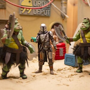 K&ouml;nnte beinhalten: Drei Actionfiguren, zwei gr&uuml;ne und eine silberne, stehen auf einer sandigen Oberfl&auml;che. Die gr&uuml;nen Figuren sind humanoid mit gro&szlig;en K&ouml;pfen und tragen braune Lederg&uuml;rtel und schwarze R&ouml;cke. Die silberne Figur ist ein Mandalorianer-Krieger mit einem Helm und einem Blastergewehr. Die Figuren sind in einer konfrontativen Haltung aufgestellt.