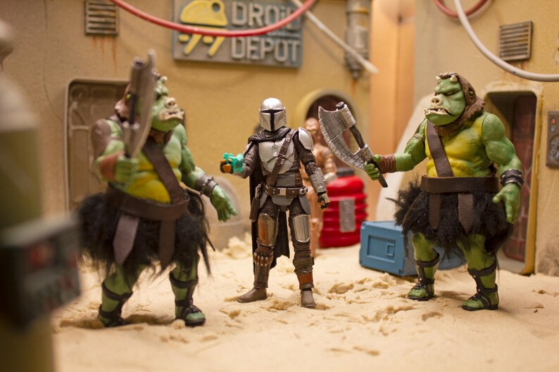 K&ouml;nnte beinhalten: Drei Actionfiguren, zwei gr&uuml;ne und eine silberne, stehen auf einer sandigen Oberfl&auml;che. Die gr&uuml;nen Figuren sind humanoid mit gro&szlig;en K&ouml;pfen und tragen braune Lederg&uuml;rtel und schwarze R&ouml;cke. Die silberne Figur ist ein Mandalorianer-Krieger mit einem Helm und einem Blastergewehr. Die Figuren sind in einer konfrontativen Haltung aufgestellt.