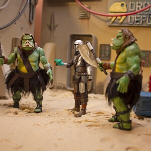 K&ouml;nnte beinhalten: Drei Actionfiguren, zwei gr&uuml;ne und eine silberne, stehen auf einer sandigen Oberfl&auml;che. Die gr&uuml;nen Figuren sind humanoid mit gro&szlig;en K&ouml;pfen und halten Waffen. Die silberne Figur tr&auml;gt eine R&uuml;stung und h&auml;lt eine Waffe. Die Figuren stehen vor einem Hintergrund eines Geb&auml;udes mit dem Text "DROID DEPOT" darauf.