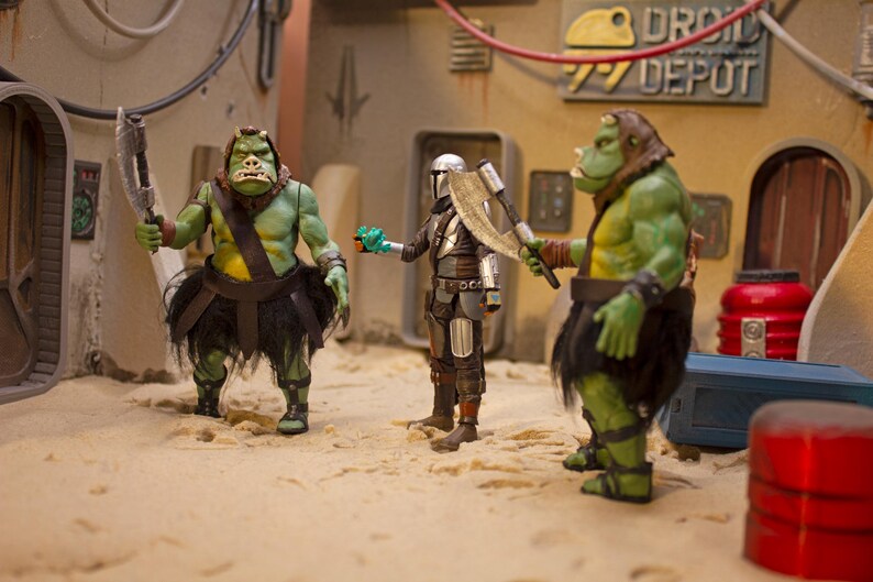 K&ouml;nnte beinhalten: Drei Actionfiguren, zwei gr&uuml;ne und eine silberne, stehen auf einer sandigen Oberfl&auml;che. Die gr&uuml;nen Figuren sind humanoid mit gro&szlig;en K&ouml;pfen und halten Waffen. Die silberne Figur tr&auml;gt eine R&uuml;stung und h&auml;lt eine Waffe. Die Figuren stehen vor einem Hintergrund eines Geb&auml;udes mit dem Text "DROID DEPOT" darauf.