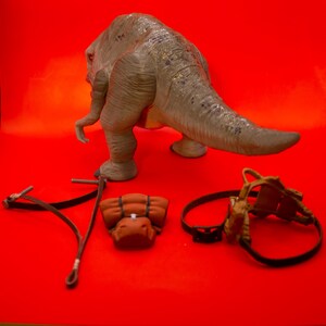 Puede incluir: Un juguete de dinosaurio gris con una cola larga, una mochila marr&oacute;n, una correa de cuero marr&oacute;n y un arn&eacute;s de cuero marr&oacute;n sobre un fondo rojo.