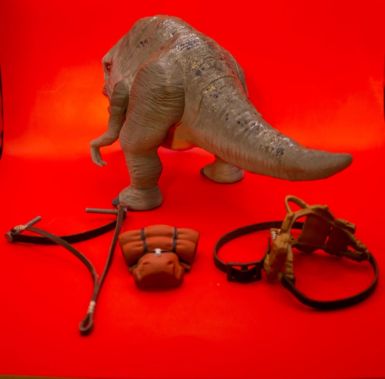 Puede incluir: Un juguete de dinosaurio gris con una cola larga, una mochila marr&oacute;n, una correa de cuero marr&oacute;n y un arn&eacute;s de cuero marr&oacute;n sobre un fondo rojo.