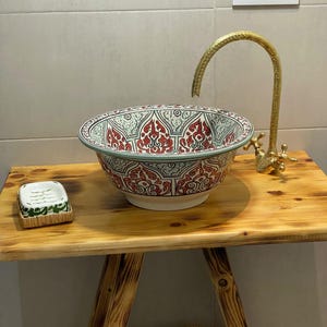 May include: A patterned ceramic sink with red, white, and green designs. The sink is on a wooden table with a gold tap. A small soap dish is on the table. The sink is a unique bathroom fixture.