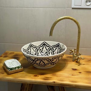 May include: A decorative bathroom sink with a black and white patterned design. A gold faucet and a small soap dish sit on a wooden table. The sink is a vessel style, and the faucet has a curved spout.