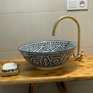 Può includere: Un lavabo decorativo in ceramica con un motivo blu e bianco. Il lavabo è montato su un supporto in legno con un rubinetto dorato. Una saponetta poggia sul supporto. Il lavabo è largo circa 46 cm.