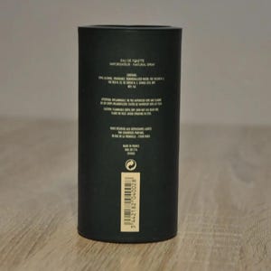 May include: Black cylindrical bottle with a gold label. The label reads "Eau de Toilette Vaporisateur Natural Spray". The bottle is on a wooden surface.