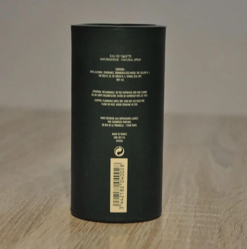 May include: Black cylindrical bottle with a gold label. The label reads "Eau de Toilette Vaporisateur Natural Spray". The bottle is on a wooden surface.
