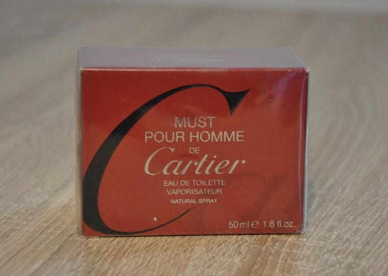 May include: A red and black Cartier Must de Cartier Pour Homme Eau de Toilette Natural Spray perfume box. The box features the iconic Cartier logo and text indicating the fragrance name, type, and volume (50ml/1.6 fl.oz.). The packaging is sealed in clear plastic wrap, suggesting a new, unopened product. The image showcases a classic men's fragrance in its original retail packaging.