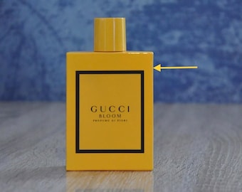 May include: A yellow rectangular perfume bottle with a matching cap. The bottle has a black border around a label that reads "GUCCI BLOOM PROFUMO DI FIORI". The background is a blurred blue and the bottle sits on a wooden surface.