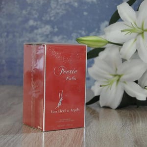 Può includere: Una scatola rossa con un'etichetta bianca che dice "Feerie Rubis" e "Van Cleef & Arpels" con una piccola illustrazione di un fiore. La scatola è per un profumo da 100 ml / 3,3 fl oz.