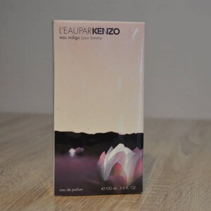 Op de afbeelding: Een rechthoekige parfumdoos met de tekst "L'EAUPARKENZO eau indigo pour femme" in het zwart. De doos heeft een lichtroze achtergrond met een donkerpaars en wit bloemmotief aan de onderkant. Bevat 100 ml - 3,4 FL. OZ.