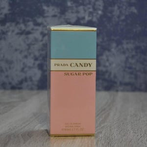 Peut inclure: Boîte de parfum rectangulaire avec un dessus bleu clair et un bas rose. Le texte "PRADA CANDY SUGAR POP" est imprimé sur le devant. La boîte a une bordure dorée et le bas indique "e 80ml 2.7 FL OZ".