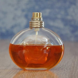 Pu&ograve; includere: Flacone di profumo rotondo e trasparente riempito di liquido color ambra. Il flacone ha un tappo dorato e un vaporizzatore. Lo sfondo &egrave; blu e bianco sfocato. Su una superficie di legno.