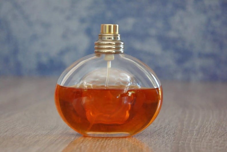 Pu&ograve; includere: Flacone di profumo rotondo e trasparente riempito di liquido color ambra. Il flacone ha un tappo dorato e un vaporizzatore. Lo sfondo &egrave; blu e bianco sfocato. Su una superficie di legno.