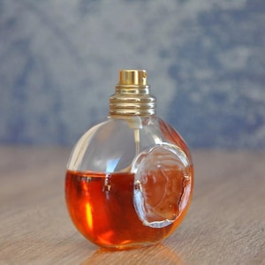 Pu&ograve; includere: Un flacone di profumo trasparente e rotondo con un tappo color oro e un design in rilievo. Il flacone &egrave; riempito con un liquido rosso-arancio. Lo sfondo &egrave; blu e bianco sfocato.
