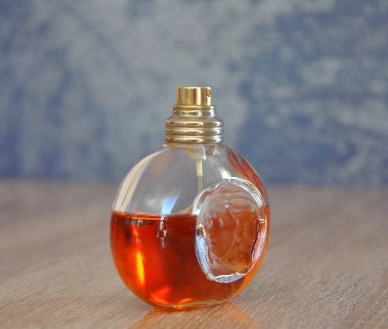 Pu&ograve; includere: Un flacone di profumo trasparente e rotondo con un tappo color oro e un design in rilievo. Il flacone &egrave; riempito con un liquido rosso-arancio. Lo sfondo &egrave; blu e bianco sfocato.