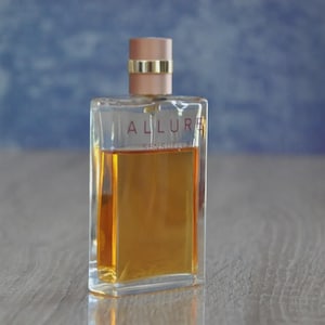May include: A rectangular perfume bottle with a clear glass body and a light brown liquid inside. The bottle has a light pink cap with a gold band. The word "ALLURE" is printed on the front of the bottle.
