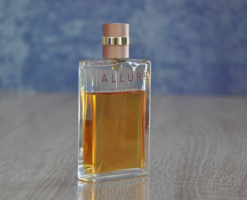 May include: A rectangular perfume bottle with a clear glass body and a light brown liquid inside. The bottle has a light pink cap with a gold band. The word "ALLURE" is printed on the front of the bottle.