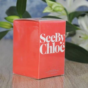 May include: A rectangular, coral-colored box with the words "See By Chlo&eacute;" in white lettering. The box is set against a backdrop of white flowers and green foliage, suggesting a fragrance product.