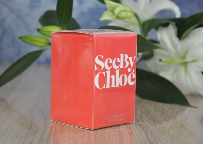 May include: A rectangular, coral-colored box with the words "See By Chlo&eacute;" in white lettering. The box is set against a backdrop of white flowers and green foliage, suggesting a fragrance product.