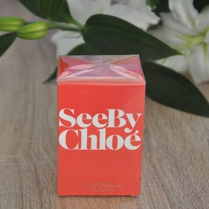 May include: A rectangular, red box of See By Chlo&eacute; perfume. The brand name is in white, bold letters. The box is cellophane-wrapped. White flowers and green leaves are in the background.