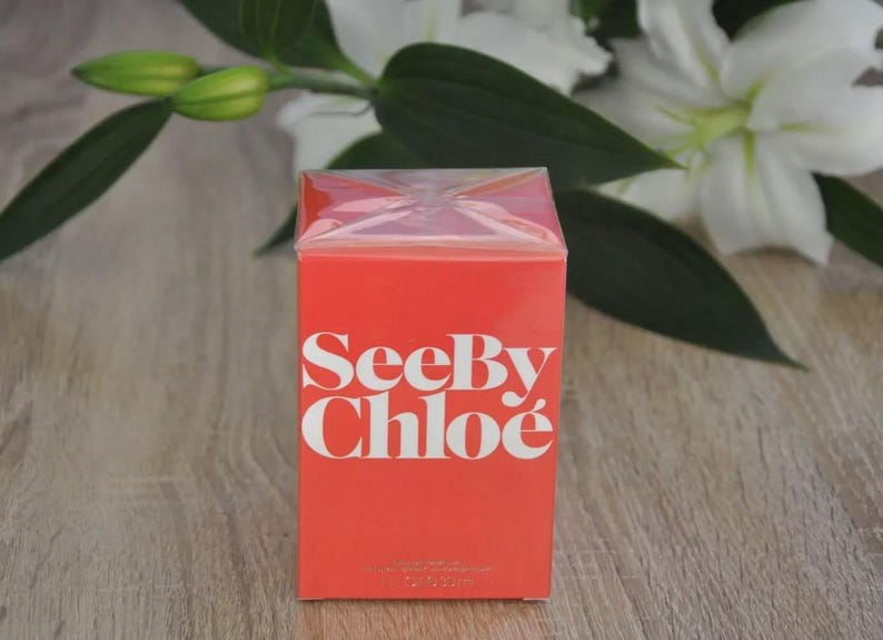 May include: A rectangular, red box of See By Chlo&eacute; perfume. The brand name is in white, bold letters. The box is cellophane-wrapped. White flowers and green leaves are in the background.