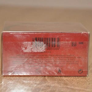 May include: Red product box with a barcode and other product information printed on it.  The box is sealed in plastic wrap. The barcode number is 005564.  There is also a small QR code and other text indicating product details and warnings. The box appears to contain a cosmetic or fragrance product.