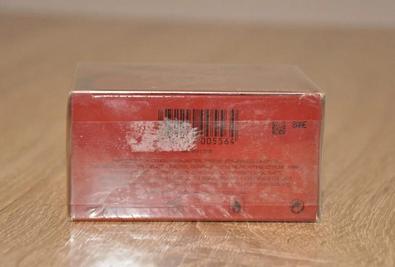 May include: Red product box with a barcode and other product information printed on it.  The box is sealed in plastic wrap. The barcode number is 005564.  There is also a small QR code and other text indicating product details and warnings. The box appears to contain a cosmetic or fragrance product.