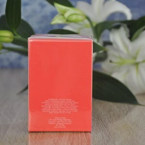 May include: A rectangular, coral-colored product box with white text, set against a backdrop of white lilies and green foliage. The box has a glossy finish and appears to contain a fragrance or cosmetic product.