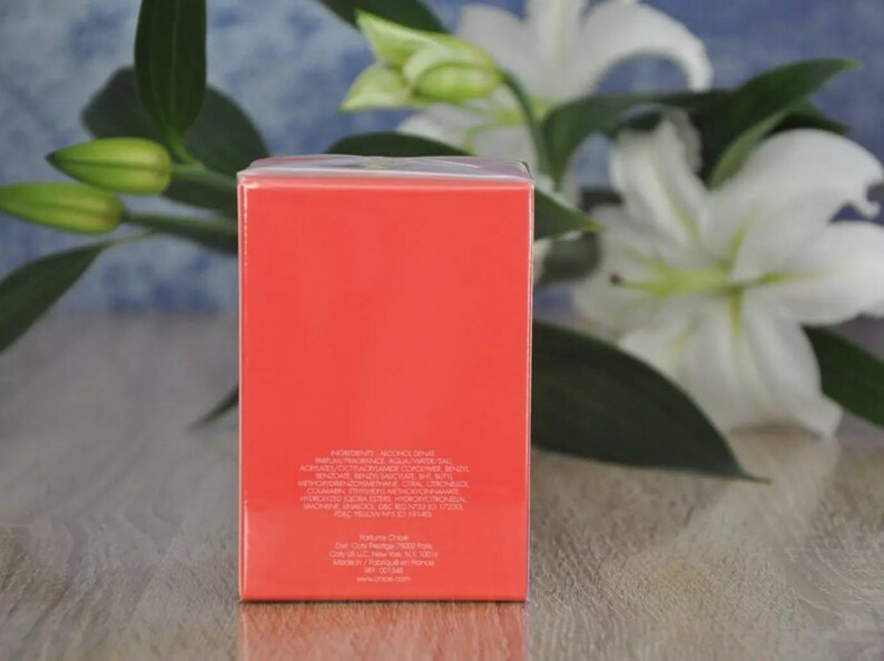 May include: A rectangular, coral-colored product box with white text, set against a backdrop of white lilies and green foliage. The box has a glossy finish and appears to contain a fragrance or cosmetic product.
