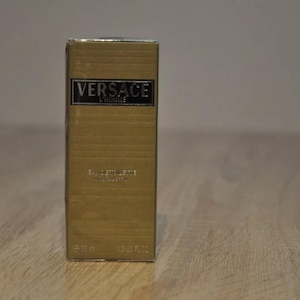 Może przedstawiać: Złote pudełko z nadrukiem "VERSACE L'HOMME" na przodzie. Pudełko jest do męskiego zapachu.