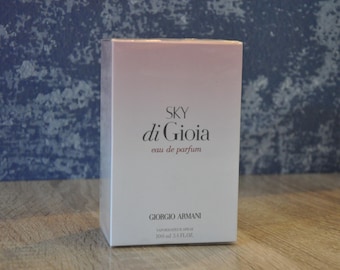 May include: A rectangular perfume box with a gradient pink and white design. The box features the text "SKY di Gioia eau de parfum" and the Giorgio Armani logo. The box is sealed in clear plastic and is on a wooden surface.