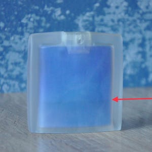 Puede incluir: Un frasco de perfume transparente y rectangular con líquido azul en su interior. El frasco tiene un aspecto esmerilado y una forma cuadrada. El fondo es azul borroso. El frasco está sobre una superficie de madera.
