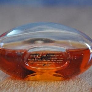 Pu&ograve; includere: Flacone di profumo trasparente, di forma ovale, riempito con un liquido marrone-rossastro. Il flacone ha una parte superiore arrotondata e una base piatta. Contiene 100 ml. Il flacone &egrave; su una superficie di legno.