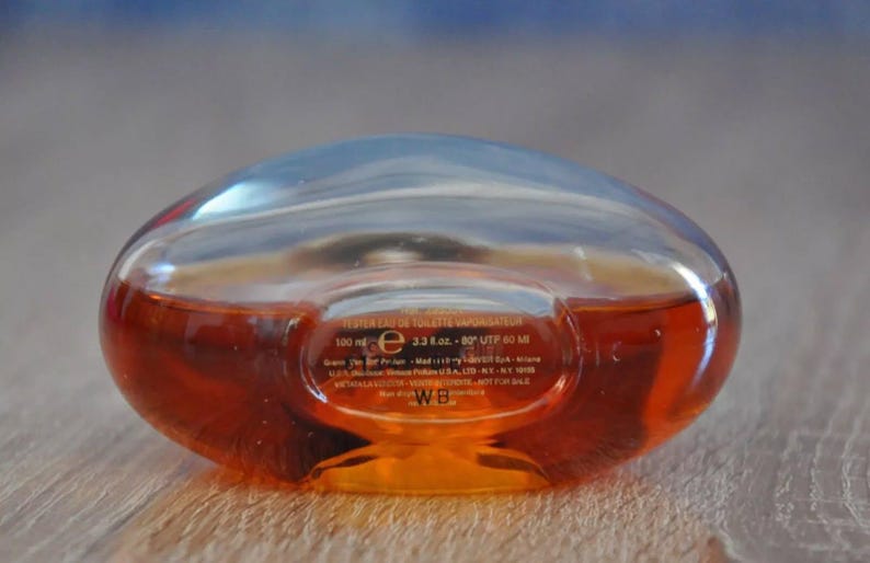 Pu&ograve; includere: Flacone di profumo trasparente, di forma ovale, riempito con un liquido marrone-rossastro. Il flacone ha una parte superiore arrotondata e una base piatta. Contiene 100 ml. Il flacone &egrave; su una superficie di legno.