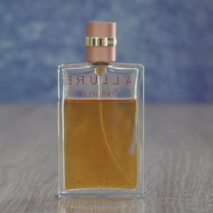 May include: A rectangular perfume bottle with a clear glass body and a light brown liquid inside. The bottle has a rose gold cap and the word "ALLURE" is printed on the front. The background is a blurred blue and white.