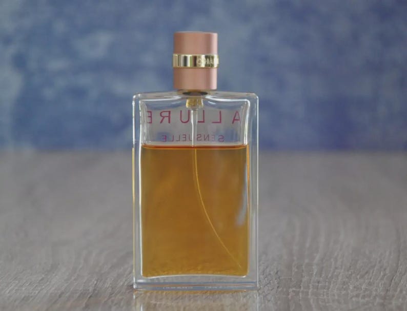 May include: A rectangular perfume bottle with a clear glass body and a light brown liquid inside. The bottle has a rose gold cap and the word "ALLURE" is printed on the front. The background is a blurred blue and white.
