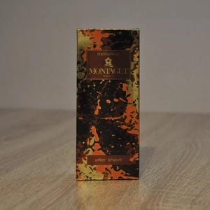 May include: A gold and black aftershave bottle with a floral design and the text "monsieur MONTAGUT PARIS" on the front. The bottle is on a wooden surface.